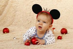 portret van schattig weinig meisje met muis oren Aan een beige plaid en rood glimmend Kerstmis ballen. foto