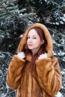 jong mooi vrouw met rood haar- en blauw ogen in faux vacht jas is wandelen in winter besneeuwd park. foto