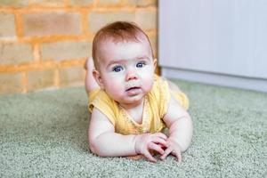 portret van glimlachen weinig Kaukasisch baby meisje in geel jurk met houten tandjes krijgen speelgoed. kind proberen naar kruipen Aan een vloer. foto