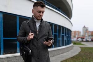 jong Mens met een mobiel telefoon en een bedrijf zak Aan de achtergrond van een parkeren veel foto