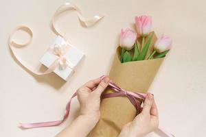 vlak vrouw handen stropdas een satijn lint boog Aan een gemakkelijk boeket van vers roze tulp bloemen in ambacht papier Aan een beige tafel. foto