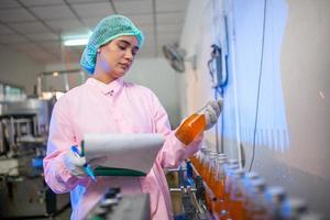 Aziatisch vrouw arbeider met klembord is controle Product flessen van fruit sap Aan de productie lijn in de drank fabriek. fabrikant cheques kwaliteit van voedsel industrie. foto