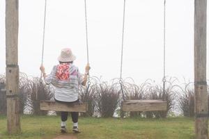 achterzijde visie van senior vrouw zittend alleen Aan schommels in tuin Aan nevelig dag foto