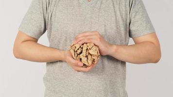 de man's hand- is verscheuren verfrommeld bruin papier bal Aan wit achtergrond. hij draagt een grijs t-shirt. foto