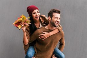 genieten van herfst tijd samen. knap jong Mens meeliften zijn vriendin Holding boeket van oranje gedaald bladeren met grijs muur net zo achtergrond foto