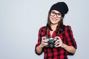 elegant fotograaf. mooi jong vrouw in hoofddeksels en bril Holding camera en op zoek Bij camera terwijl staand tegen grijs achtergrond foto