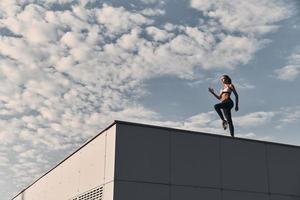 macht en schoonheid. vol lengte van modern jong vrouw in sport- kleding jumping terwijl oefenen tijd Aan de dak buitenshuis foto