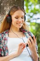 genieten van muziek- en vers lucht. mooi jong vrouw in hoofdtelefoons luisteren naar de muziek- en glimlachen terwijl leunend Bij de boom in een park foto