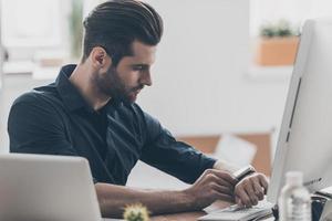 zelfverzekerd Mens. knap jong het professioneel gebruik makend van slim kijk maar terwijl zittend Bij de bureau in creatief kantoor foto