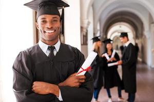 zelfverzekerd afstuderen. gelukkig Afrikaanse Mens in diploma uitreiking jurken Holding diploma en glimlachen terwijl zijn vrienden staand in de achtergrond foto