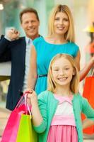 boodschappen doen samen is plezier. vrolijk familie Holding boodschappen doen Tassen en glimlachen Bij camera terwijl staand in boodschappen doen winkelcentrum foto