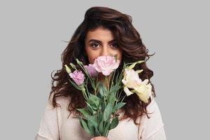 mooi net zo een bloem. aantrekkelijk jong vrouw aan het bedekken gezicht met bundel van bloemen en op zoek Bij camera terwijl staand tegen grijs achtergrond foto