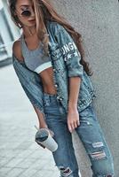 gewoontjes stijl. aantrekkelijk jong vrouw in jeans slijtage Holding een beschikbaar kop terwijl leunend Aan de bouwkundig kolom buitenshuis foto