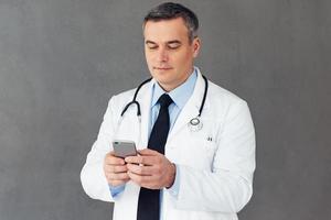 op zoek door zijn rooster. volwassen mannetje dokter gebruik makend van zijn slim telefoon terwijl staand tegen grijs achtergrond foto