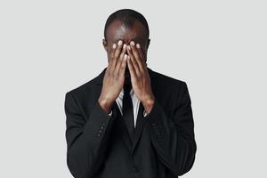 gefrustreerd jong Afrikaanse Mens in formele kleding aan het bedekken gezicht met handen terwijl staand tegen grijs achtergrond foto