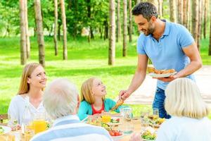 genieten van tijd met familie. gelukkig familie van vijf mensen genieten van maaltijd samen terwijl zittend Bij de dining tafel in formeel tuin foto