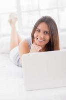 surfing de netto Bij huis. mooi jong glimlachen vrouw aan het liegen in bed en gebruik makend van laptop foto