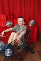 senior elegant grijs haren vrouw in zonnebril en zilver jurk zittend in de supermarkt kar Bij de partij, drinken wijn Aan rood gordijnen achtergrond. partij, disco, viering, senior leeftijd concept foto