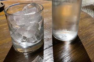 selectief focus, een Doorzichtig glas gevulde met ijs en Doorzichtig glas fles houdt verkoudheid mineraal water zit Aan houten tafel foto