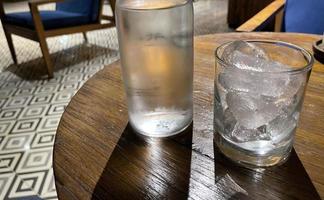 selectief focus, een Doorzichtig glas gevulde met ijs en Doorzichtig glas fles houdt verkoudheid mineraal water zit Aan houten tafel foto