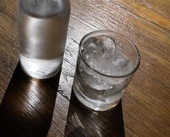 selectief focus, een Doorzichtig glas gevulde met ijs en Doorzichtig glas fles houdt verkoudheid mineraal water zit Aan houten tafel foto