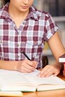 aan het doen haar huiswerk. jong vrouw schrijven iets in haar Notitie stootkussen en lezing boek terwijl zittend Bij de bureau foto