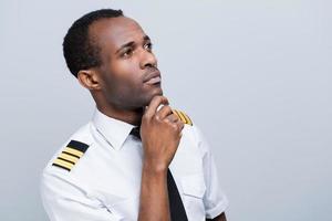 attent piloot. kant visie van attent Afrikaanse piloot in uniform Holding hand- Aan kin terwijl staand tegen grijs achtergrond foto