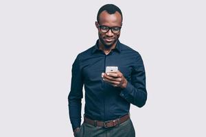 typen snel bericht. knap jong Afrikaanse Mens Holding slim telefoon en op zoek Bij het terwijl staand tegen grijs achtergrond foto