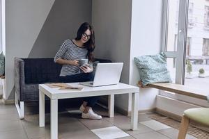 genieten van koffie Bij haar werken plaats. mooi jong vrouw Holding een kop en een slim telefoon terwijl zittend Aan de sofa foto