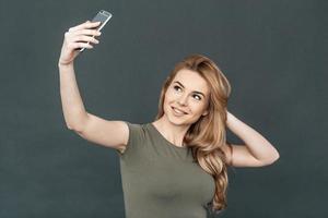 nemen selfie. portret van aantrekkelijk jong glimlachen vrouw met blond haar- maken selfie door haar slim telefoon terwijl staand tegen grijs achtergrond foto