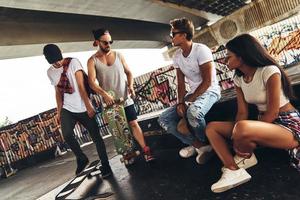 nemen een pauze. groep van jong modern mensen pratend terwijl uitgeven tijd Bij de skateboard park buitenshuis foto