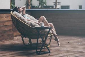 uitgeven zorgeloos tijd. mooi jong vrouw ontspannende in een groot comfortabel stoel Aan haar buitenshuis huis terras foto