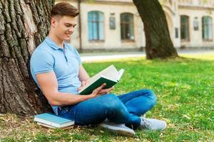 lezing zijn favoriete boek. zelfverzekerd mannetje leerling lezing boek terwijl zittend Aan de gras en in voorkant van Universiteit gebouw foto