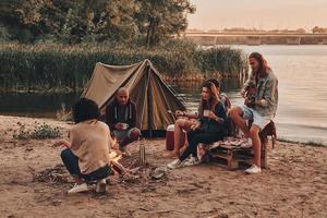 momenten van vreugde. groep van jong mensen in gewoontjes slijtage glimlachen terwijl genieten van strand partij in de buurt de kampvuur foto