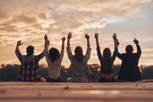 vriendschap dat duurt voor altijd. achterzijde visie van jong mensen in gewoontjes slijtage roosteren met een bier flessen terwijl zittend Aan de pier foto