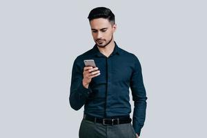 typen snel bericht. echt jong Mens in overhemd typen tekst bericht gebruik makend van zijn slim telefoon terwijl staand tegen grijs achtergrond foto