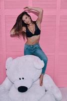 gevoel speels. aantrekkelijk jong vrouw spelen met groot speelgoed- beer en glimlachen terwijl staand tegen roze achtergrond foto