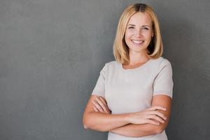 zelfverzekerd schoonheid. vrolijk volwassen vrouw houden armen gekruiste en glimlachen terwijl staand tegen grijs achtergrond foto