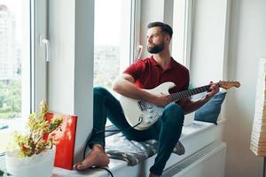romantisch jong Mens in gewoontjes kleding spelen gitaar terwijl zittend Aan de venster dorpel foto