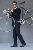 elegant en stijlvol. vol lengte van zelfverzekerd jong zakenman draag- zijn fiets Aan schouder terwijl staand tegen grijs achtergrond foto