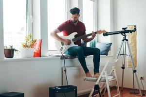 knap jong Mens in gewoontjes kleding spelen gitaar terwijl zittend Aan de venster dorpel foto