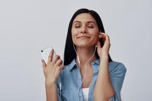 aantrekkelijk jong vrouw luisteren muziek- en glimlachen terwijl staand tegen grijs achtergrond foto