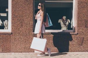 na dag winkelen. kant visie van mooi jong vrouw draag- boodschappen doen Tassen en glimlachen terwijl wandelen langs de straat met mode op te slaan in de achtergrond foto