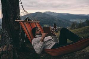 vertrekken allemaal uw zorgen achter. knap jong Mens aan het liegen in hangmat en lezing een boek terwijl camping met zijn vriendin foto