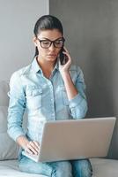 belangrijk bedrijf telefoongesprek. mooi jong vrouw in bril pratend Aan mobiel telefoon en werken Aan laptop terwijl zittend Aan de bankstel tegen grijs achtergrond foto