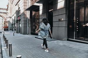 straat stijl. vol lengte achterzijde visie van jong vrouw in denim jasje wandelen naar beneden de straat foto