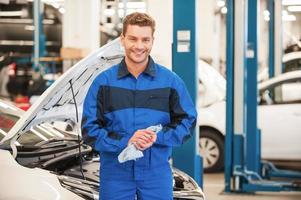 na de werk is gedaan. zelfverzekerd jong Mens in uniform afvegen zijn handen met vod en glimlachen terwijl staand in werkplaats met auto in de achtergrond foto