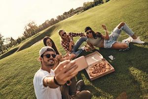 creëren gelukkig herinneringen. groep van jong glimlachen mensen in gewoontjes slijtage nemen selfie terwijl genieten van voedsel en drankjes buitenshuis foto