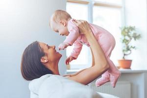 u zijn mijn schat kant visie van vrolijk mooi jong vrouw Holding baby meisje in haar handen en op zoek Bij haar met liefde terwijl zittend Aan de bankstel Bij huis foto