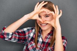 liefde u mooi jong vrouw maken hart vorm met haar handen en op zoek Bij camera met glimlach terwijl staand tegen grijs achtergrond foto